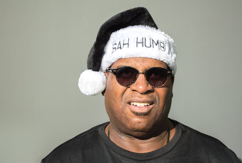 Cranky middle aged rotund African American man wearing a black and white Santa hat saying Bah Humbug against a solid background. Cranky middle aged rotund African American man wearing a black and white Santa hat saying Bah Humbug against a solid background