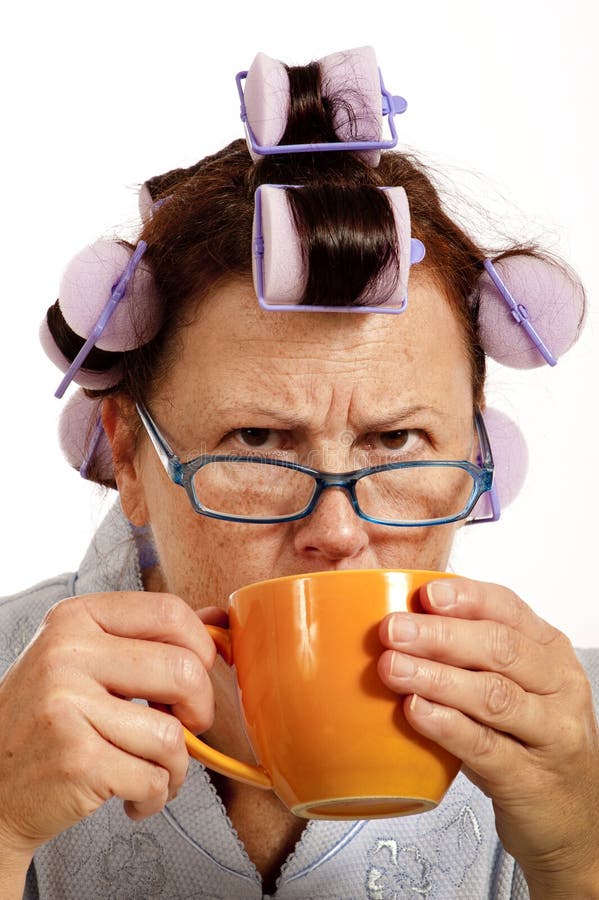 Vertical shot of a mature grumpy woman in curlers drinking coffee in the morning. On white background