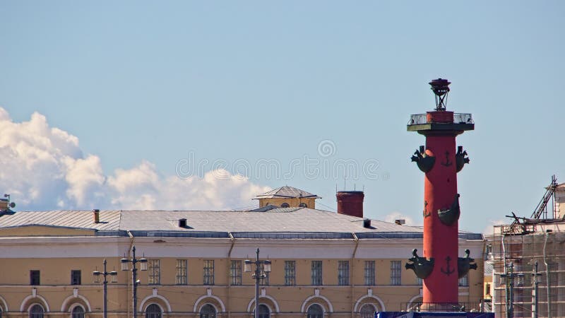 View of a fragment of an ancient building on the embankment and a huge red rostral column with stylized bows of ancient ships as decoration and a large gas torch on top, one of the symbols of the city, ancient traditions, history and culture, sky and clouds, cityscape, summer. View of a fragment of an ancient building on the embankment and a huge red rostral column with stylized bows of ancient ships as decoration and a large gas torch on top, one of the symbols of the city, ancient traditions, history and culture, sky and clouds, cityscape, summer.