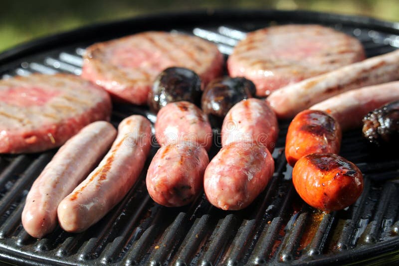 Grunt fokusskott av smakliga blandade läckra korvar och hamburgare som fräser och lagar mat på en grillfestlaggplatta, yttersida royaltyfria bilder Grunt fokusskott av smakliga blandade läckra korvar och hamburgare som fräser och lagar mat på en grillfestlaggplatta, yttersida royaltyfria bilder