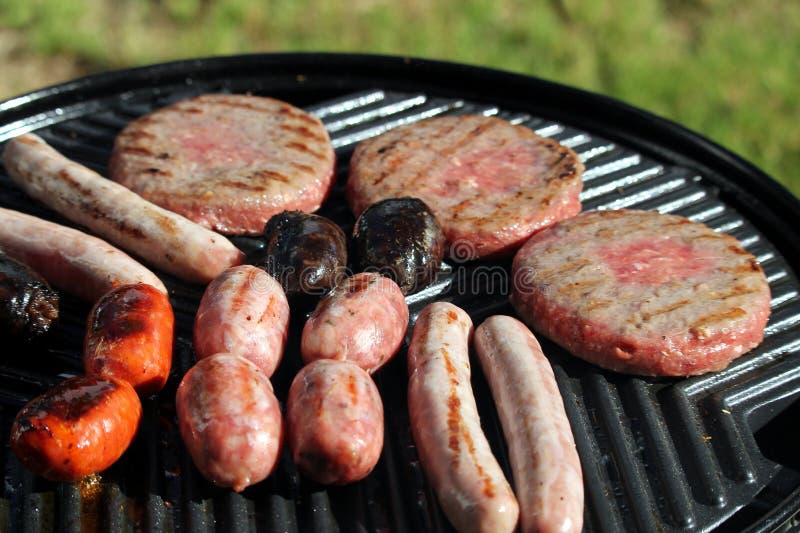 Grunt fokusskott av smakliga blandade läckra korvar och hamburgare som fräser och lagar mat på en grillfestlaggplatta, yttersida royaltyfria bilder Grunt fokusskott av smakliga blandade läckra korvar och hamburgare som fräser och lagar mat på en grillfestlaggplatta, yttersida royaltyfria bilder