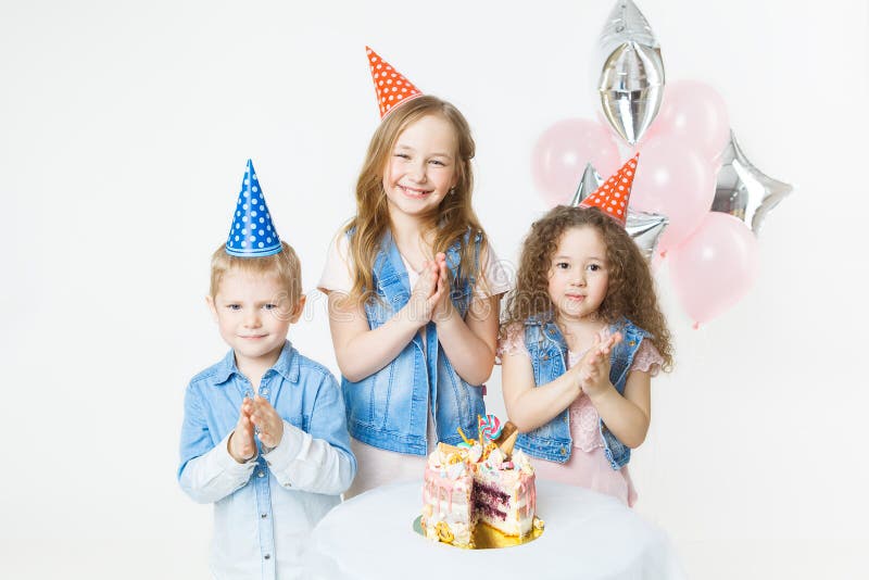 Group of kids in festive caps clap their hands near birthday cake, balloons on background. Studio. Group of kids in festive caps clap their hands near birthday cake, balloons on background. Studio