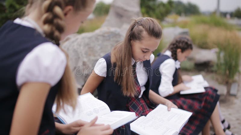 Grupa siedzi outdoors i opowiada młodzi przyjaciół ucznie jest ubranym ten sam mundurek szkolnego podczas gdy czytający książkę w