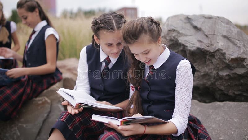 Grupa siedzi outdoors i opowiada młodzi przyjaciół ucznie jest ubranym ten sam mundurek szkolnego podczas gdy czytający książkę d