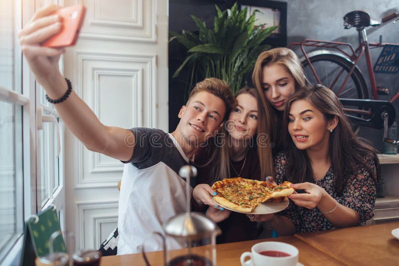 Grupo de adolescentes lindos que toman el selfie con el teléfono móvil mientras que se sienta en un restaurante con el interior e imagen de archivo libre de regalías Grupo de adolescentes lindos que toman el selfie con el teléfono móvil mientras que se sienta en un restaurante con el interior e imagen de archivo libre de regalías