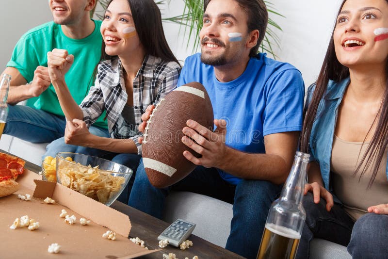 Grupo de aficionados desportivos dos amigos que olha o fósforo no futebol americano das camisas coloridas imagens de stock Grupo de aficionados desportivos dos amigos que olha o fósforo no futebol americano das camisas coloridas imagens de stock
