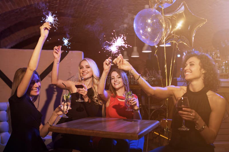 Grupo de amigos fêmeas que apreciam a festa de anos que tem o divertimento com chuveirinhos do fogo de artifício que bebe o assen foto de stock Grupo de amigos fêmeas que apreciam a festa de anos que tem o divertimento com chuveirinhos do fogo de artifício que bebe o assen foto de stock