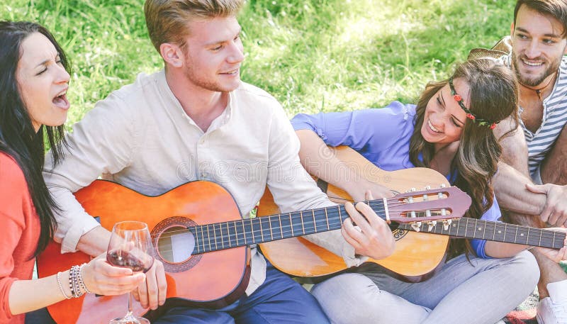 Grupo de amigos que tocan las guitarras y que cantan mientras que bebe el vino tinto que se sienta en hierba en un parque al aire imagenes de archivo Grupo de amigos que tocan las guitarras y que cantan mientras que bebe el vino tinto que se sienta en hierba en un parque al aire imagenes de archivo