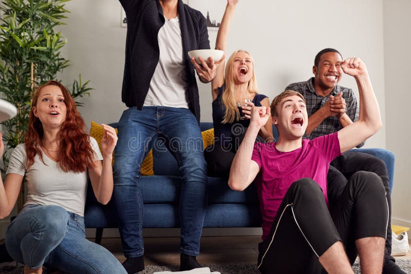 Grupo De Amigos Universitarios Celebrando Mientras Miran Juego Deportivo En Televisión En Shared House foto de archivo libre de regalías