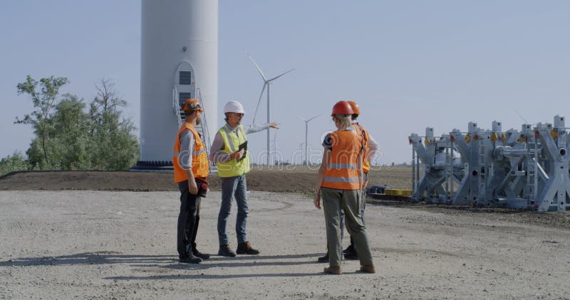 Full length colleagues in vests and helmets examining and discussing construction site of wind farm in countryside. Full length colleagues in vests and helmets examining and discussing construction site of wind farm in countryside