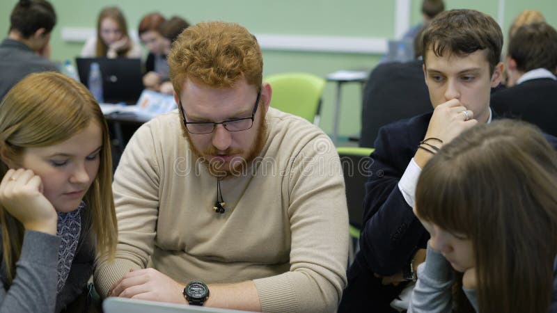 Grupo de estudiantes universitarios que se sientan junto en el escritorio en sala de clase, mirando los ordenadores y haciendo pr