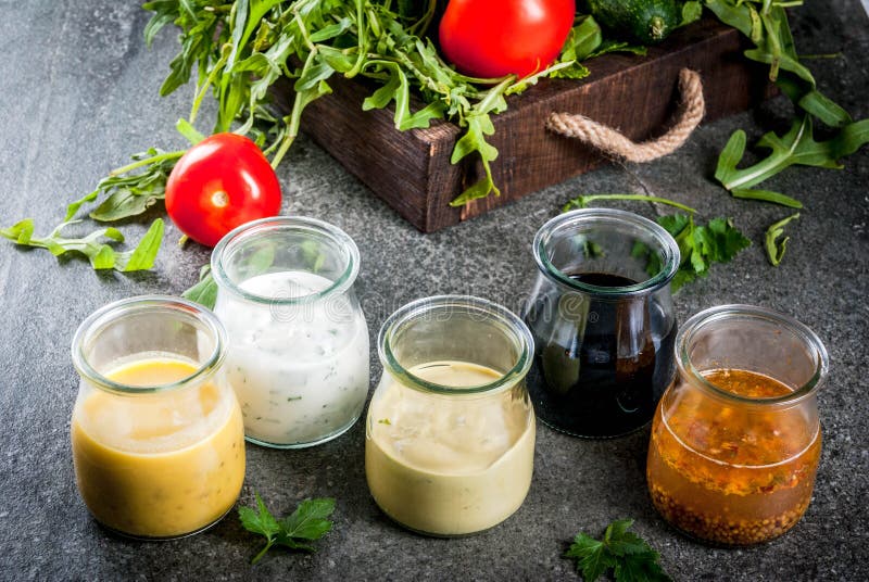 Set of dressings for salad: sauce vinaigrette, mustard, mayonnaise or ranch, balsamic or soy, basil with yogurt. Dark stone table. On background of greenery, vegetables for salad. Copy space. Set of dressings for salad: sauce vinaigrette, mustard, mayonnaise or ranch, balsamic or soy, basil with yogurt. Dark stone table. On background of greenery, vegetables for salad. Copy space