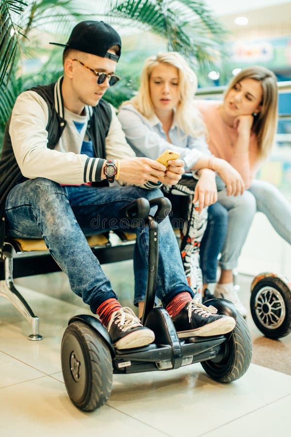 Grupo de personas en el hoverboard eléctrico de la vespa que se sienta en el banco y que usa el teléfono fotos de archivo libres de regalías Grupo de personas en el hoverboard eléctrico de la vespa que se sienta en el banco y que usa el teléfono fotos de archivo libres de regalías