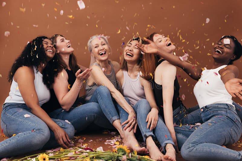Grupo de seis risas de mujeres de diferentes edades sentadas bajo pétalos de flores en caída. hembras sonrientes multiétnicas que foto de archivo