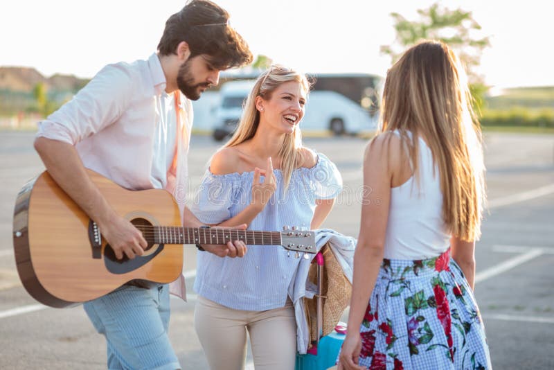 Grupo de turistas novos que têm o divertimento e que jogam a guitarra em um parque de estacionamento, transporte de espera fotografia de stock royalty free Grupo de turistas novos que têm o divertimento e que jogam a guitarra em um parque de estacionamento, transporte de espera fotografia de stock royalty free