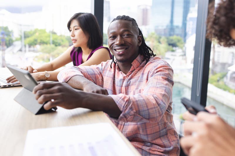 Diverse group of young adults working together in a modern business office, smiling at camera. African American men with dreadlocks wearing plaid shirt, Asian women in pink, unaltered. Diverse group of young adults working together in a modern business office, smiling at camera. African American men with dreadlocks wearing plaid shirt, Asian women in pink, unaltered