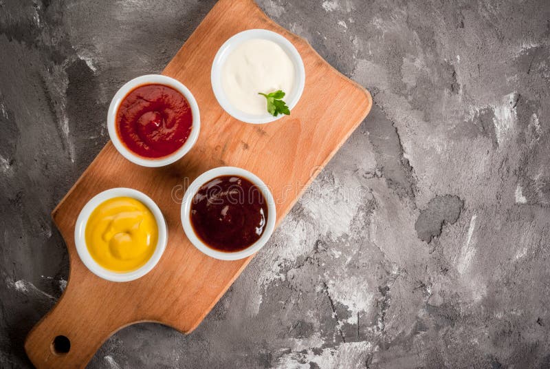Classic set of sauces in white saucers: American yellow mustard, ketchup, barbecue sauce, mayonnaise. On grey black spotted concrete stone table top view, copy space. Classic set of sauces in white saucers: American yellow mustard, ketchup, barbecue sauce, mayonnaise. On grey black spotted concrete stone table top view, copy space