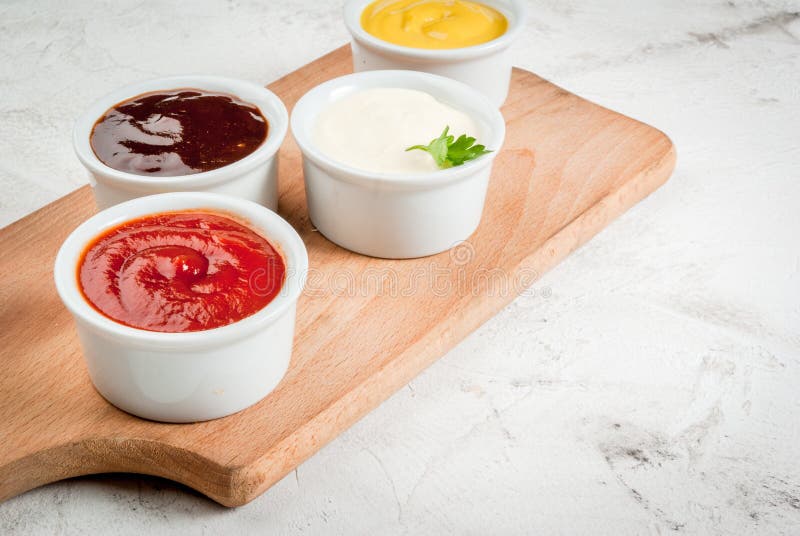 Classic set of sauces in white saucers: American yellow mustard, ketchup, barbecue sauce, mayonnaise. On cutting board white stone concrete table close view, copy space. Classic set of sauces in white saucers: American yellow mustard, ketchup, barbecue sauce, mayonnaise. On cutting board white stone concrete table close view, copy space