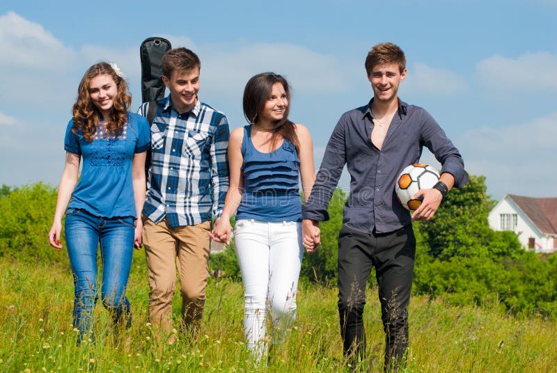 Happy mixed group of Young people two beautiful women and two handsome young men with guitar & ball walking outdoors on a bright sunny day of spring or summer on the blue sky and green field background. Happy mixed group of Young people two beautiful women and two handsome young men with guitar & ball walking outdoors on a bright sunny day of spring or summer on the blue sky and green field background