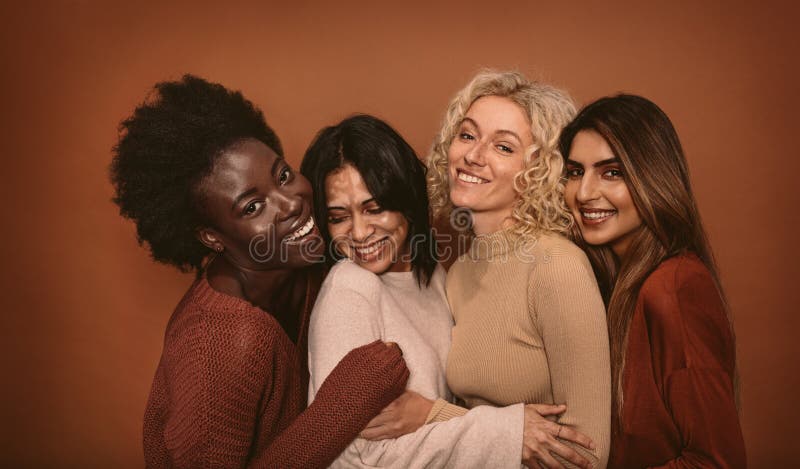 Group of cheerful young women standing together on brown background. Multi ethnic female friends in studio. Group of cheerful young women standing together on brown background. Multi ethnic female friends in studio.