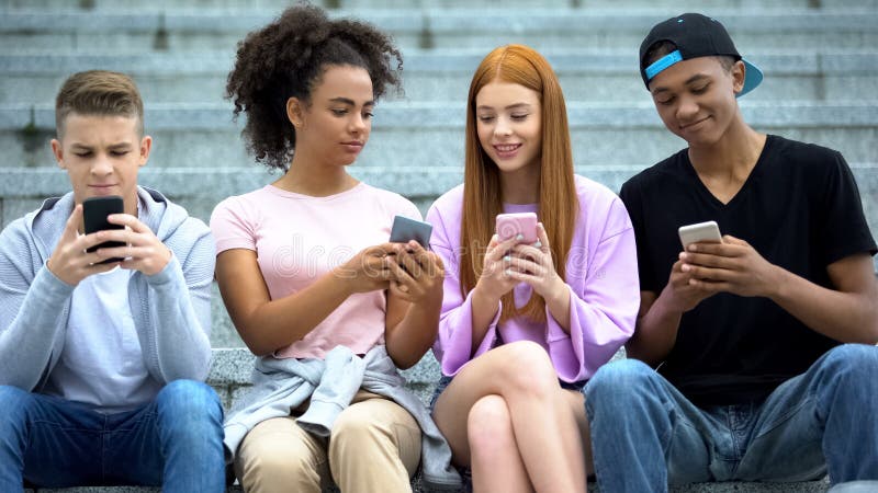 Grupp av studenter som leker smartphone-spel och sitter utomhus, gadget arkivfoto