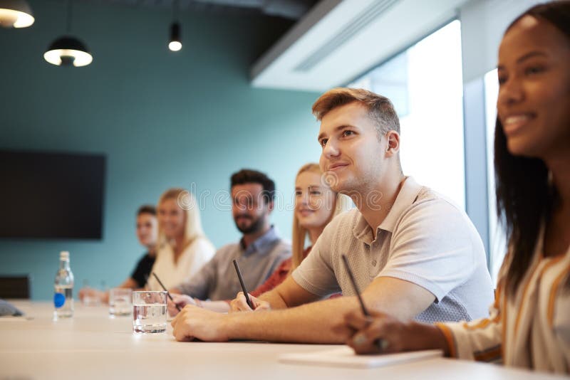 Gruppe junge Kandidaten, die am Sitzungssaal-Tisch hörend auf Darstellung Geschäfts-am graduierten Einstellungs-Einschätzungs-Tag lizenzfreie stockfotografie Gruppe junge Kandidaten, die am Sitzungssaal-Tisch hörend auf Darstellung Geschäfts-am graduierten Einstellungs-Einschätzungs-Tag lizenzfreie stockfotografie