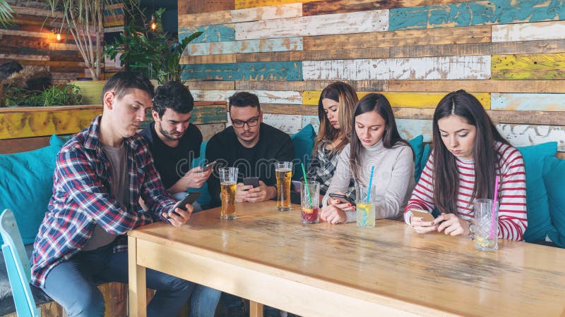 Group of friends at pub ignoring each other in favour of mobile phone, Young people addicted by mobile phone on social network community, Connected millennials interacting with smartphones always online. Group of friends at pub ignoring each other in favour of mobile phone, Young people addicted by mobile phone on social network community, Connected millennials interacting with smartphones always online
