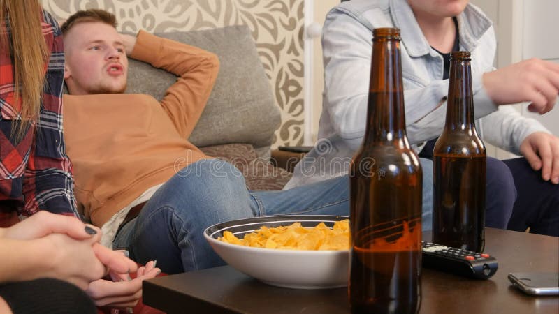 Gruppo di amici che si siedono insieme sul sofà, sulla TV di sorveglianza e sulla birra bevente fotografie stock libere da diritti Gruppo di amici che si siedono insieme sul sofà, sulla TV di sorveglianza e sulla birra bevente fotografie stock libere da diritti