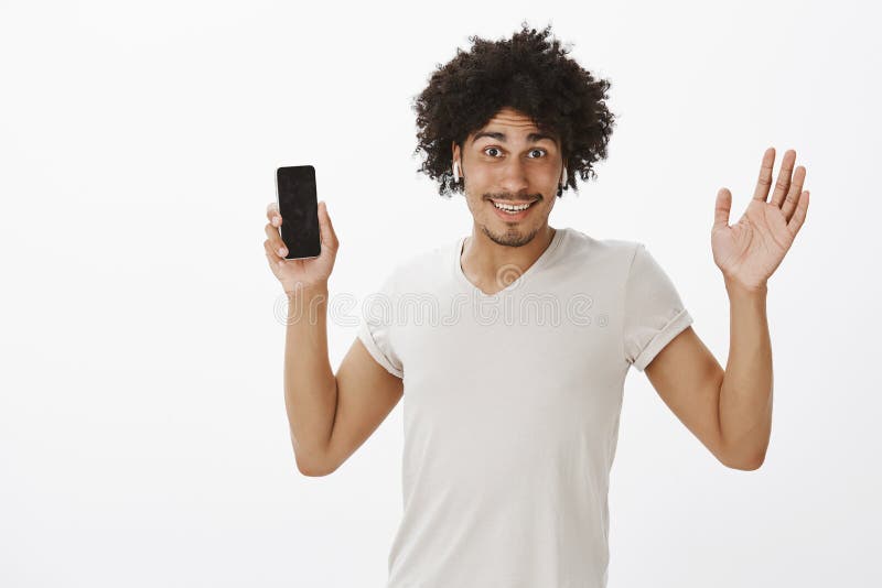 Guardi, nessun cavi Giovane maschio ispanico attraente spensierato con taglio di capelli di afro, palma di sollevamento e smartph immagine stock libera da diritti Guardi, nessun cavi Giovane maschio ispanico attraente spensierato con taglio di capelli di afro, palma di sollevamento e smartph immagine stock libera da diritti