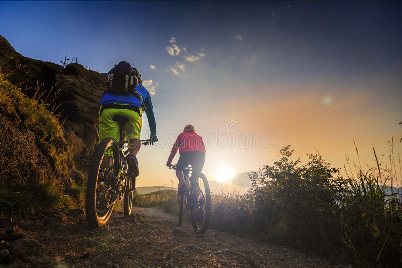 Guida delle donne e dell'uomo di ciclismo di montagna sulle bici alla montagna di tramonto
