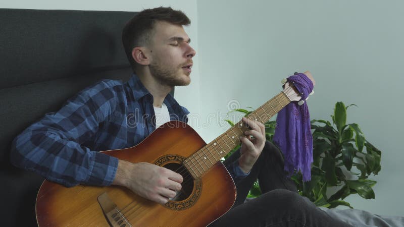 Guitar musiker spelar balladslåt på akustisk gitarr och sjunger känslomässigt hemma på sängen.