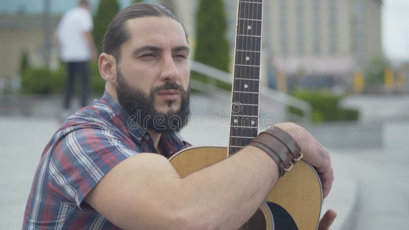 Guitarista cáucaso bonito sentado na rua urbana e olhando para o outro lado Retrato de um jovem e confiante e pensativo homem com