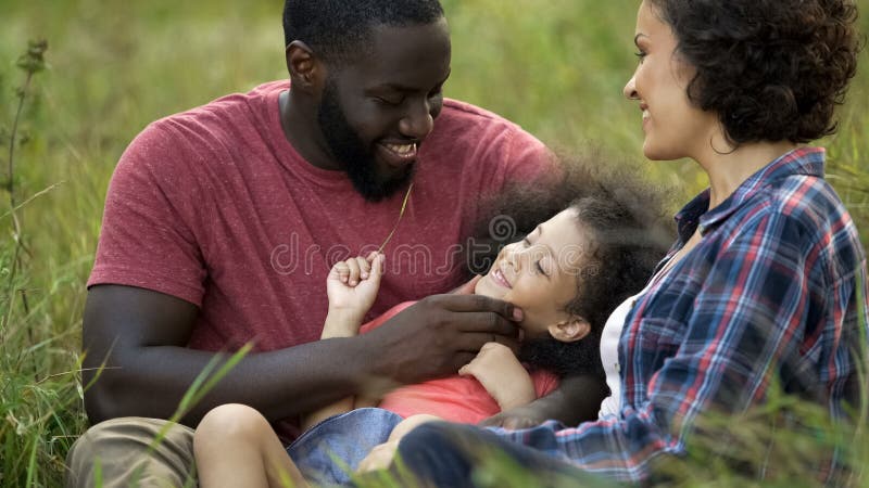 Gullig dotter som ligger i högt gräs som spelar med att bry sig föräldrar som älskar familjen royaltyfri bild Gullig dotter som ligger i högt gräs som spelar med att bry sig föräldrar som älskar familjen royaltyfri bild