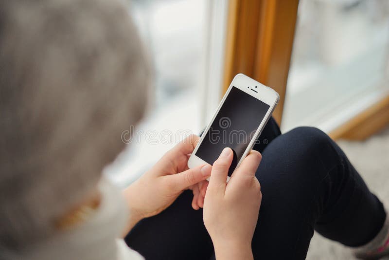 Gullig flicka med långt hår som sitter det ensamma near fönstret med mobiltelefonen royaltyfria bilder Gullig flicka med långt hår som sitter det ensamma near fönstret med mobiltelefonen royaltyfria bilder