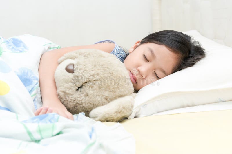 Gullig liten asiatisk björn för flickasömn- och kramnalle på säng royaltyfri foto Gullig liten asiatisk björn för flickasömn- och kramnalle på säng royaltyfri foto