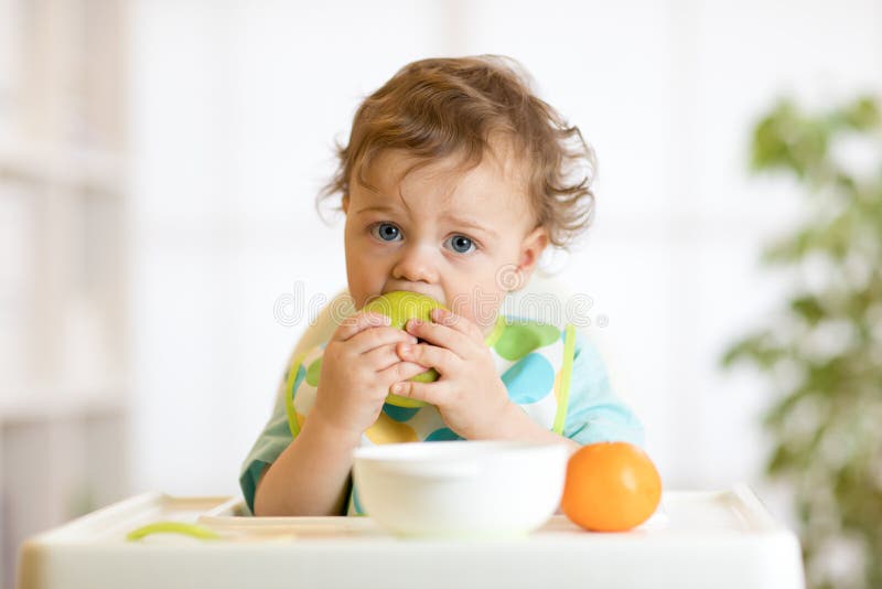 Cute baby boy one years old sitting on high children chair and eating fruits alone in white kitchen. Cute baby boy one years old sitting on high children chair and eating fruits alone in white kitchen