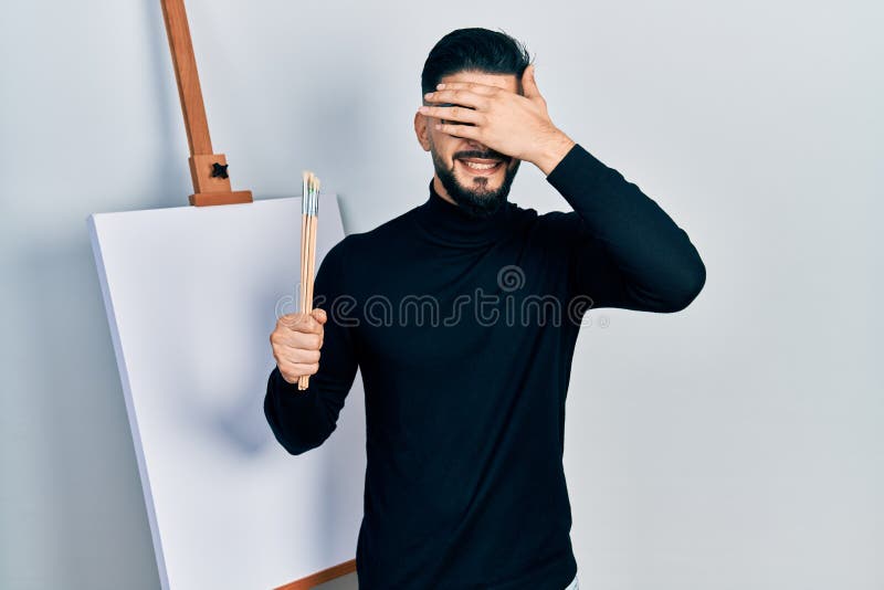Handsome man with beard holding brushes close to easel stand smiling and laughing with hand on face covering eyes for surprise. blind concept. Handsome man with beard holding brushes close to easel stand smiling and laughing with hand on face covering eyes for surprise. blind concept