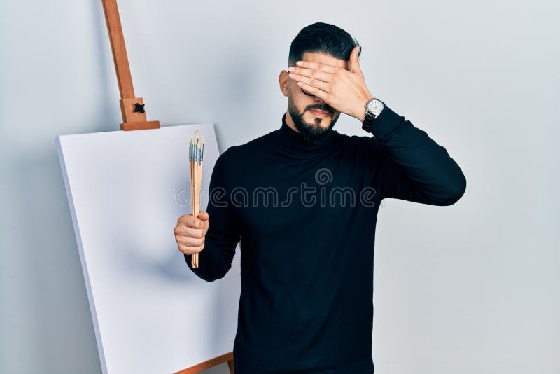 Handsome man with beard holding brushes close to easel stand covering eyes with hand, looking serious and sad. sightless, hiding and rejection concept. Handsome man with beard holding brushes close to easel stand covering eyes with hand, looking serious and sad. sightless, hiding and rejection concept