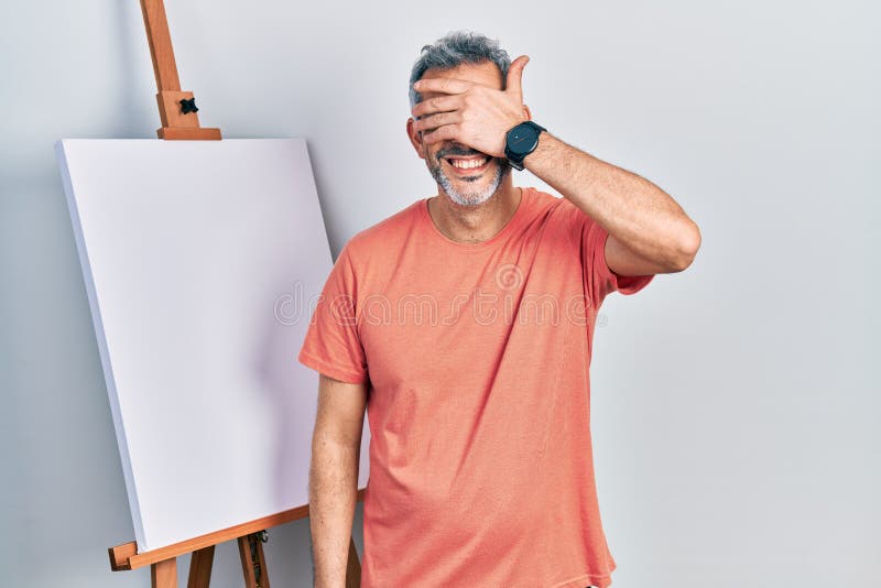 Handsome middle age man with grey hair standing by painter easel stand smiling and laughing with hand on face covering eyes for surprise. blind concept. Handsome middle age man with grey hair standing by painter easel stand smiling and laughing with hand on face covering eyes for surprise. blind concept