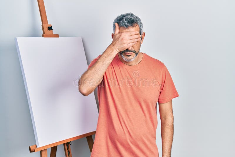 Handsome middle age man with grey hair standing by painter easel stand covering eyes with hand, looking serious and sad. sightless, hiding and rejection concept. Handsome middle age man with grey hair standing by painter easel stand covering eyes with hand, looking serious and sad. sightless, hiding and rejection concept