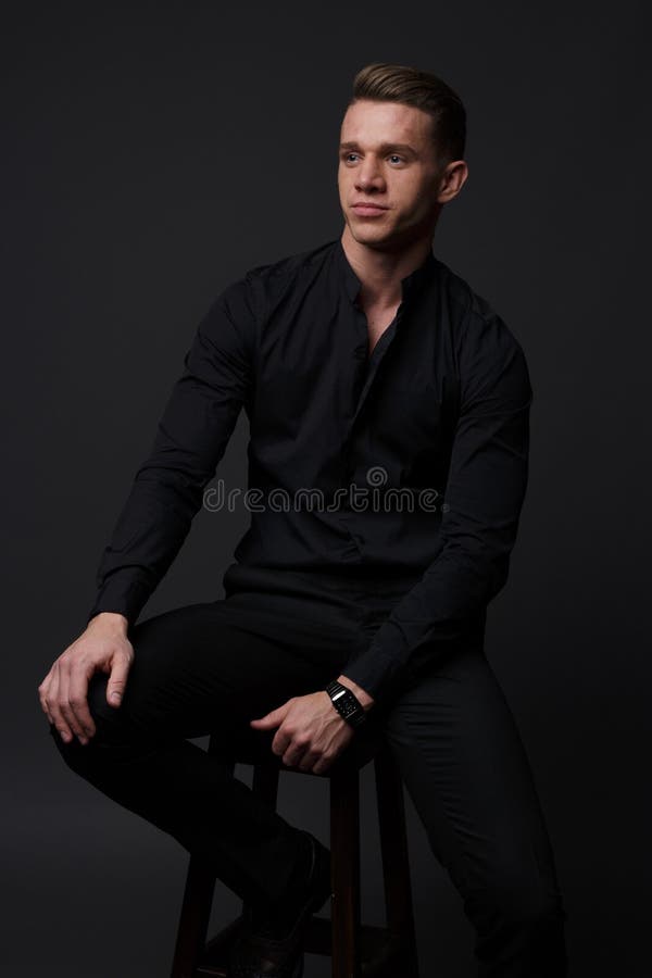 a guy in a black shirt and black pants sits on a dark chair, on a gray background