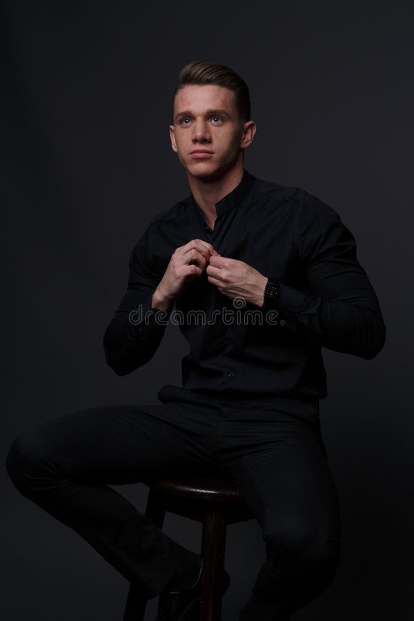 a guy in a black shirt and black pants sits on a dark chair, on a gray background