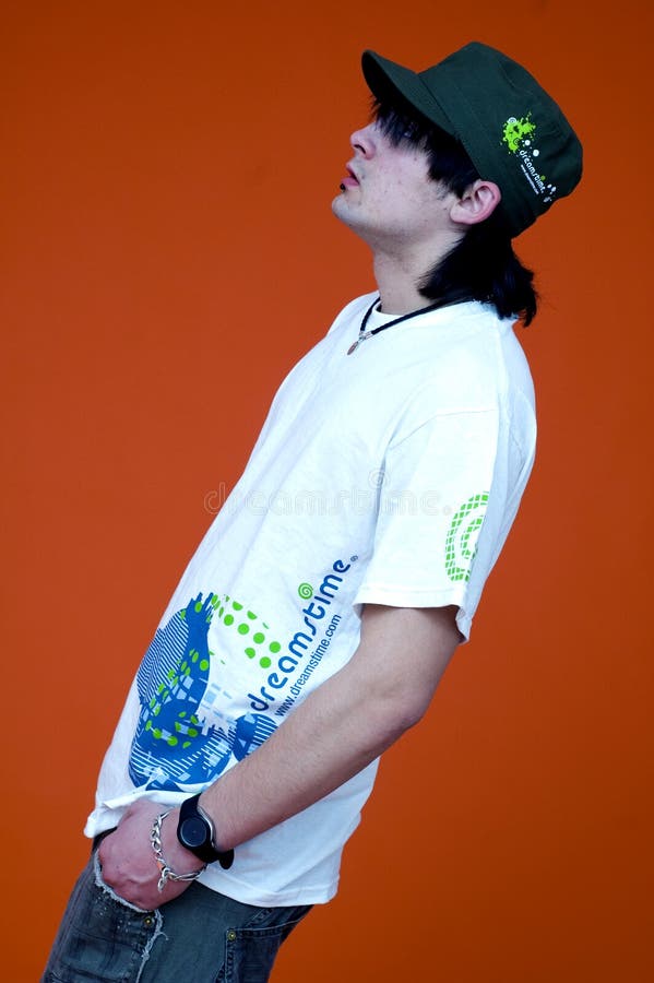 Young man or teenager with long hair wearing dreamstime shirt and cap, orange background, hand in pocket. Young man or teenager with long hair wearing dreamstime shirt and cap, orange background, hand in pocket