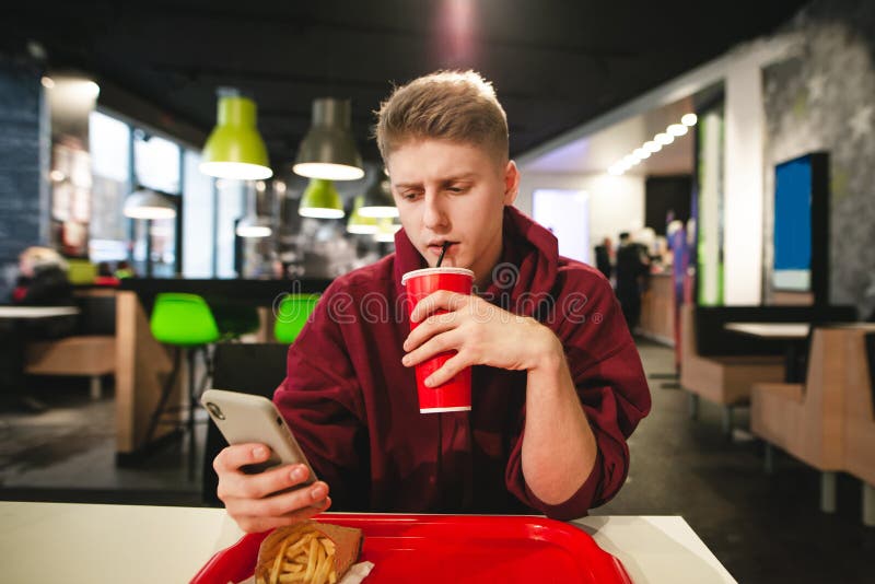 Guy drinks a beverage from a red glass and uses a smartphone in a fast food cafe stock photo Guy drinks a beverage from a red glass and uses a smartphone in a fast food cafe stock photo