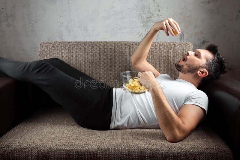 The guy in the shirt is lying on the couch, eating chips and watching a sports channel. The concept of laziness