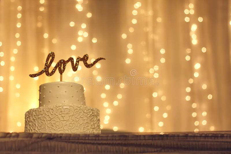 A simple white wedding cake with the word love written in sparkling gold letters on the top, with white twinkling lights and fabric in the background. A simple white wedding cake with the word love written in sparkling gold letters on the top, with white twinkling lights and fabric in the background