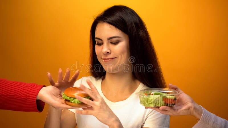 Głodna dziewczyna wybiera hamburger zamiast sałatki, tani szybkie żarcie vs zdrowa dieta zdjęcie royalty free Głodna dziewczyna wybiera hamburger zamiast sałatki, tani szybkie żarcie vs zdrowa dieta zdjęcie royalty free