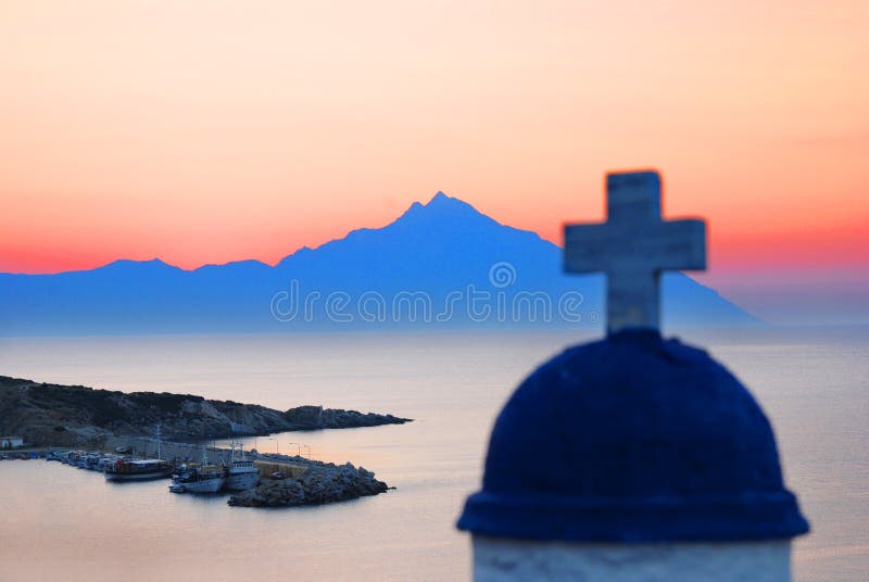 A beautiful blue silhouette of Mount Athos across the bay from Chalkidiki, Greece at sunrise with the top of a church steeple in the foreground. A beautiful blue silhouette of Mount Athos across the bay from Chalkidiki, Greece at sunrise with the top of a church steeple in the foreground.