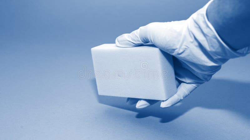 Banner with human hand in white glove holds a white melamine household sponge for cleaning on blue background toned in trendy classic blue color of year 2020. Banner with human hand in white glove holds a white melamine household sponge for cleaning on blue background toned in trendy classic blue color of year 2020.