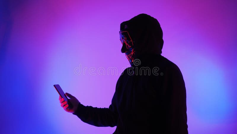 Anonymous hacker and face mask with smartphone in hand. Man in black hood shirt trying to hack personal data from mobile phone. Represent cyber crime data hacking or personal data stealing concept. Anonymous hacker and face mask with smartphone in hand. Man in black hood shirt trying to hack personal data from mobile phone. Represent cyber crime data hacking or personal data stealing concept